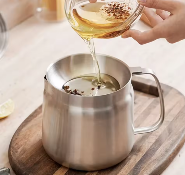 Stainless steel tea infuser with oil being poured over it on a wooden surface.