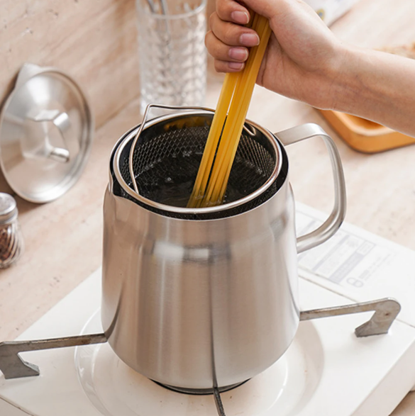 Stainless steel milk frothing pitcher with a hand holding a wooden spoon over it on a kitchen counter.